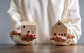 Person in a white shirt holding two small wooden house models on a table, symbolizing choice or comparison, with a calm tone