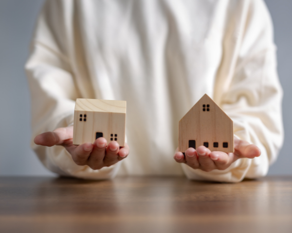 Person in a white shirt holding two small wooden house models on a table, symbolizing choice or comparison, with a calm tone
