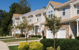 Row of modern townhouses with beige siding and stone accents under a clear blue sky. Trimmed shrubs and young trees line the driveway, creating a neat, welcoming feel.