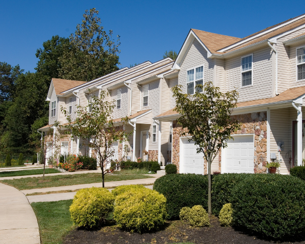 Row of modern townhouses with beige siding and stone accents under a clear blue sky. Trimmed shrubs and young trees line the driveway, creating a neat, welcoming feel.