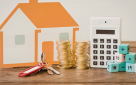 A small orange house cutout, stacks of gold coins, a calculator, house keys, and numbered cubes on a wooden surface, conveying financial planning.