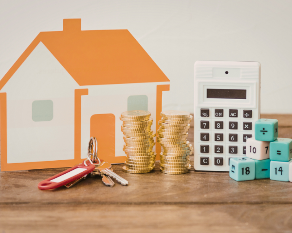 A small orange house cutout, stacks of gold coins, a calculator, house keys, and numbered cubes on a wooden surface, conveying financial planning.