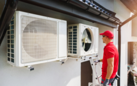 A technician in a red uniform stands on a ladder, inspecting two outdoor air conditioning units mounted on a building wall.
