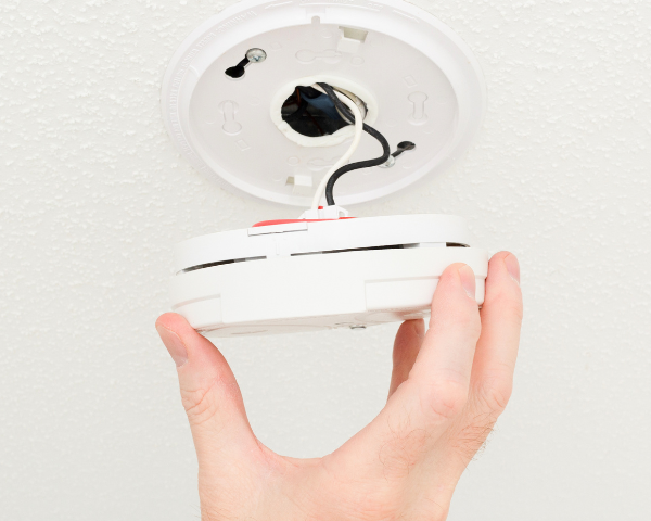 A hand is installing a smoke detector on a white ceiling. Visible are two wires and the device's base. The scene conveys attentiveness and safety.