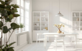 Bright, minimalist dining room with white walls and furniture. Sunlight streams through large windows, illuminating shelves and a table with a vase.