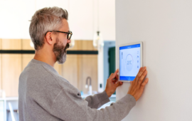Man with glasses adjusting smart thermostat on a wall in a modern home. The screen displays 23°C. The room has a calm, minimalist design.