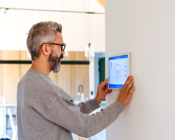 Man with glasses adjusting smart thermostat on a wall in a modern home. The screen displays 23°C. The room has a calm, minimalist design.