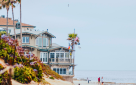 Coastal scene with a beige beach house, surrounded by palm trees and vibrant flowers. Nearby, people stroll along the sandy shore, with a calm ocean backdrop.