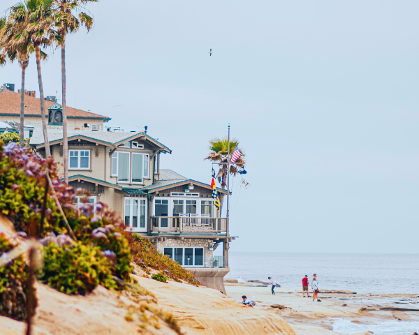 Coastal scene with a beige beach house, surrounded by palm trees and vibrant flowers. Nearby, people stroll along the sandy shore, with a calm ocean backdrop.
