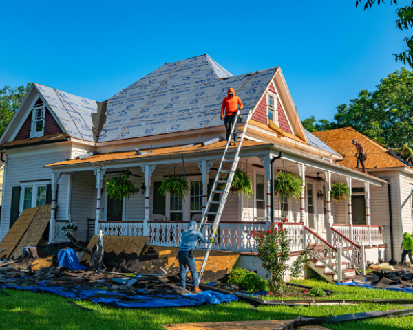 Roofers work on a house with Victorian architecture, a person climbing a ladder, surrounded by greenery and construction materials, under a clear blue sky.