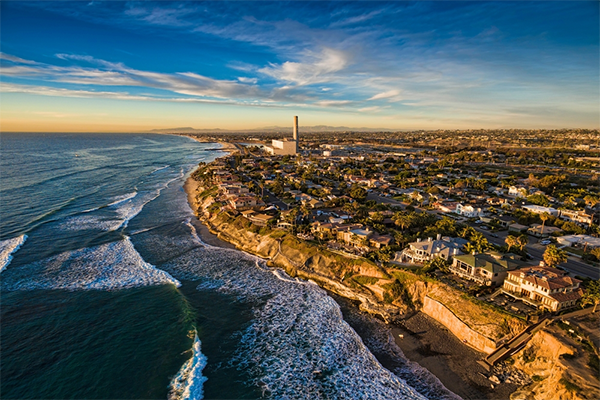 Carlsbad-Beach Aerial view of a coastal town with houses on cliffs overlooking the ocean. Waves crash against the shore, under a vibrant blue sky with wispy clouds.