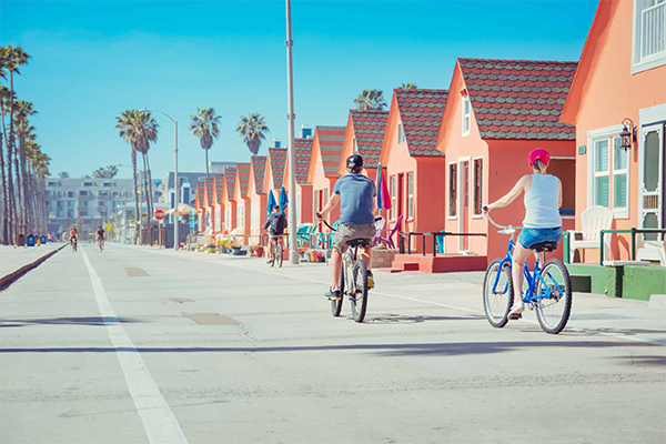 Downtown Oceanside People leisurely ride bicycles along a sunny boardwalk lined with colorful, small orange cottages and tall palm trees under a vibrant blue sky.