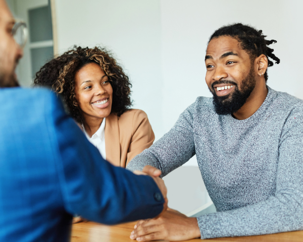 Two people, a man and a woman, smiling and sitting at a table during a meeting, while shaking hands with another person in focus, conveying a friendly, professional atmosphere.