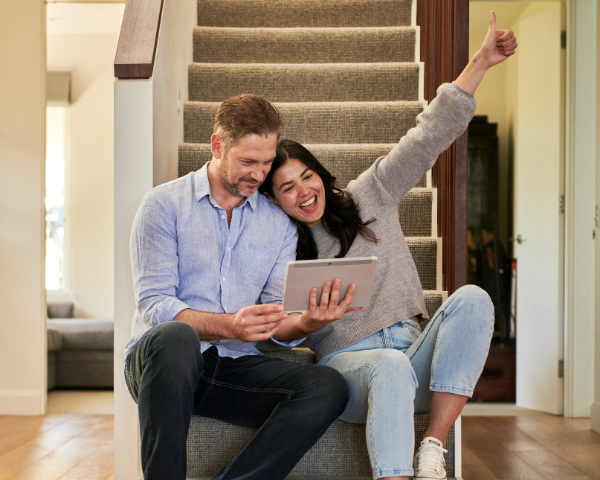A cheerful couple sits on stairs at home, the man holding a tablet and the woman raising her arm with a thumbs up, both smiling happily.