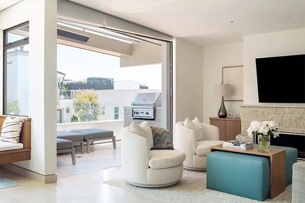 Modern living room with open patio doors showing outdoor seating and a grill. Inside, cream swivel chairs, a teal ottoman, and a mounted TV on a stone fireplace create a cozy, elegant vibe.