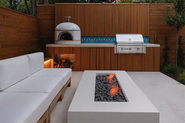 Modern patio with navy outdoor kitchen units on wheels, a black dog, and green tiled wall. Glass doors reveal an interior with orange chairs. Peaceful.
