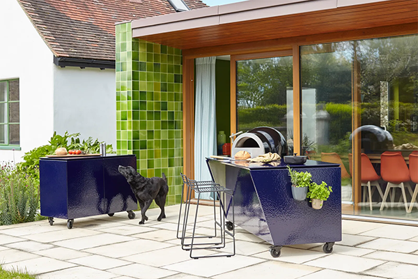 Modern patio with navy outdoor kitchen units on wheels, a black dog, and green tiled wall. Glass doors reveal an interior with orange chairs. Peaceful.