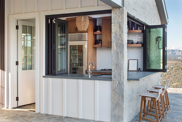 Outdoor kitchen with a modern design, featuring a stone counter, open bi-fold windows, wooden shelves, and bar stools. Scenic mountain view in the background.