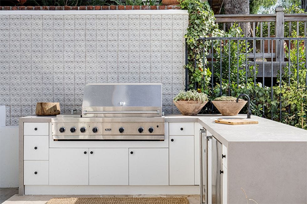 Outdoor kitchen with a stainless steel grill set in white cabinets. It has potted plants, a cutting board, and a vine-covered wall, evoking a serene, natural ambiance.