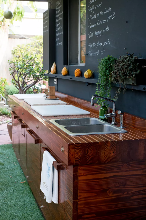 Modern patio with navy outdoor kitchen units on wheels, a black dog, and green tiled wall. Glass doors reveal an interior with orange chairs. Peaceful.