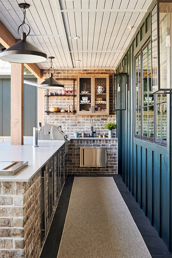 Modern patio with navy outdoor kitchen units on wheels, a black dog, and green tiled wall. Glass doors reveal an interior with orange chairs. Peaceful.