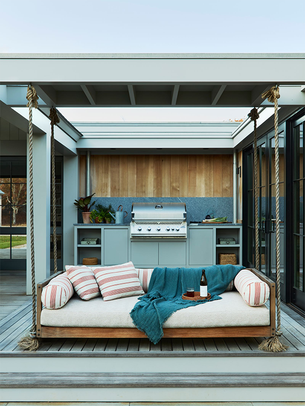 Modern patio with navy outdoor kitchen units on wheels, a black dog, and green tiled wall. Glass doors reveal an interior with orange chairs. Peaceful.