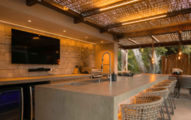 Outdoor patio with a modern kitchen. Stone bar, woven chairs, large TV, and warm lighting create a cozy, inviting atmosphere.