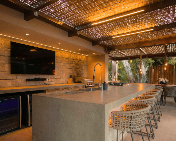 Outdoor patio with a modern kitchen. Stone bar, woven chairs, large TV, and warm lighting create a cozy, inviting atmosphere.