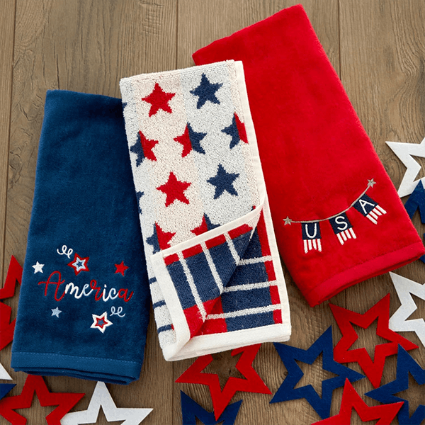 Three patriotic towels are displayed on a wooden surface. A blue towel reads _America,_ a white towel has red and blue stars, and a red towel says _USA._ Colorful star cutouts surround them. Three patriotic towels are displayed on a wooden surface. A blue towel reads _America,_ a white towel has red and blue stars, and a red towel says _USA._ Colorful star cutouts surround them.