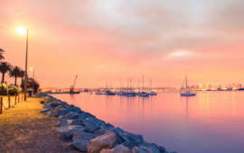 Sunset over a tranquil marina in San Diego with sailboats, casting a warm pink glow in the sky and reflecting on the water. A rocky path and palm trees are on the left.
