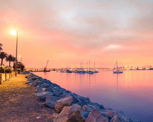 Sunset over a tranquil marina in San Diego with sailboats, casting a warm pink glow in the sky and reflecting on the water. A rocky path and palm trees are on the left.