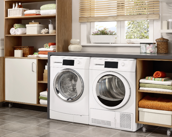 Bright, tidy laundry room with white washer and dryer, shelves of folded towels, baskets, and a sunlit window; clean, welcoming atmosphere.