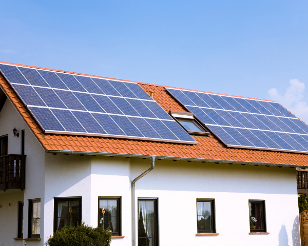 White house with a terracotta roof covered in blue solar panels beneath a clear sky, conveying clean energy and sustainable living.