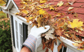 Gloved hand clears colorful autumn leaves from the gutter of a red-shingled house roof, highlighting seasonal home maintenance.