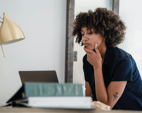 Thoughtful woman with curly hair rests her chin on her hand beside a laptop and books, appearing focused while working at home.
