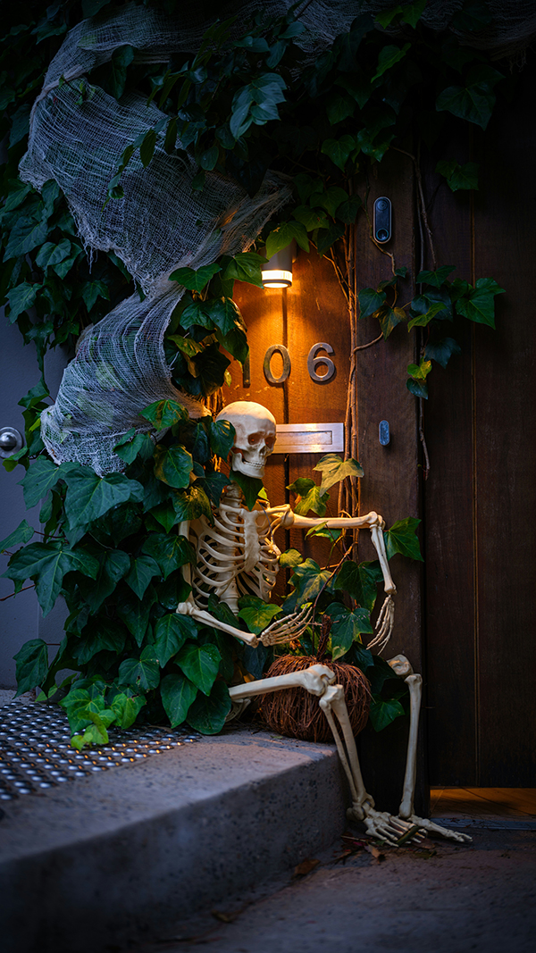 A skeleton sits beside a glowing door marked 106, tangled in ivy and cobwebs; moody Halloween decor creates a spooky, playful atmosphere.