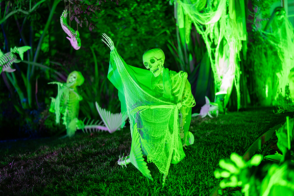 Neon-green skeletons draped in gauzy fabric stand among glowing trees and hanging decorations, creating an eerie, playful Halloween scene at night.