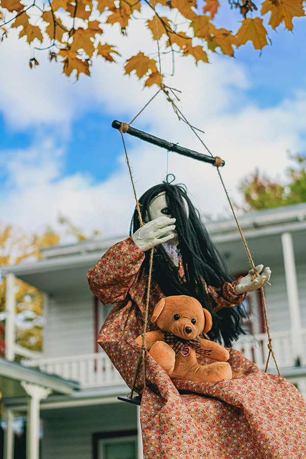 A long-haired handmade figure in a floral dress sits on a porch swing, holding a teddy bear beneath golden autumn leaves; whimsical, slightly eerie mood.