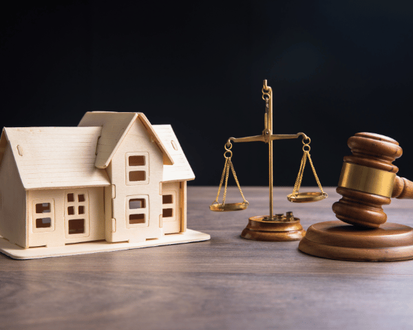 A wooden model house sits beside balanced brass scales and a judge’s gavel on a table, evoking property law and a calm, serious legal tone.